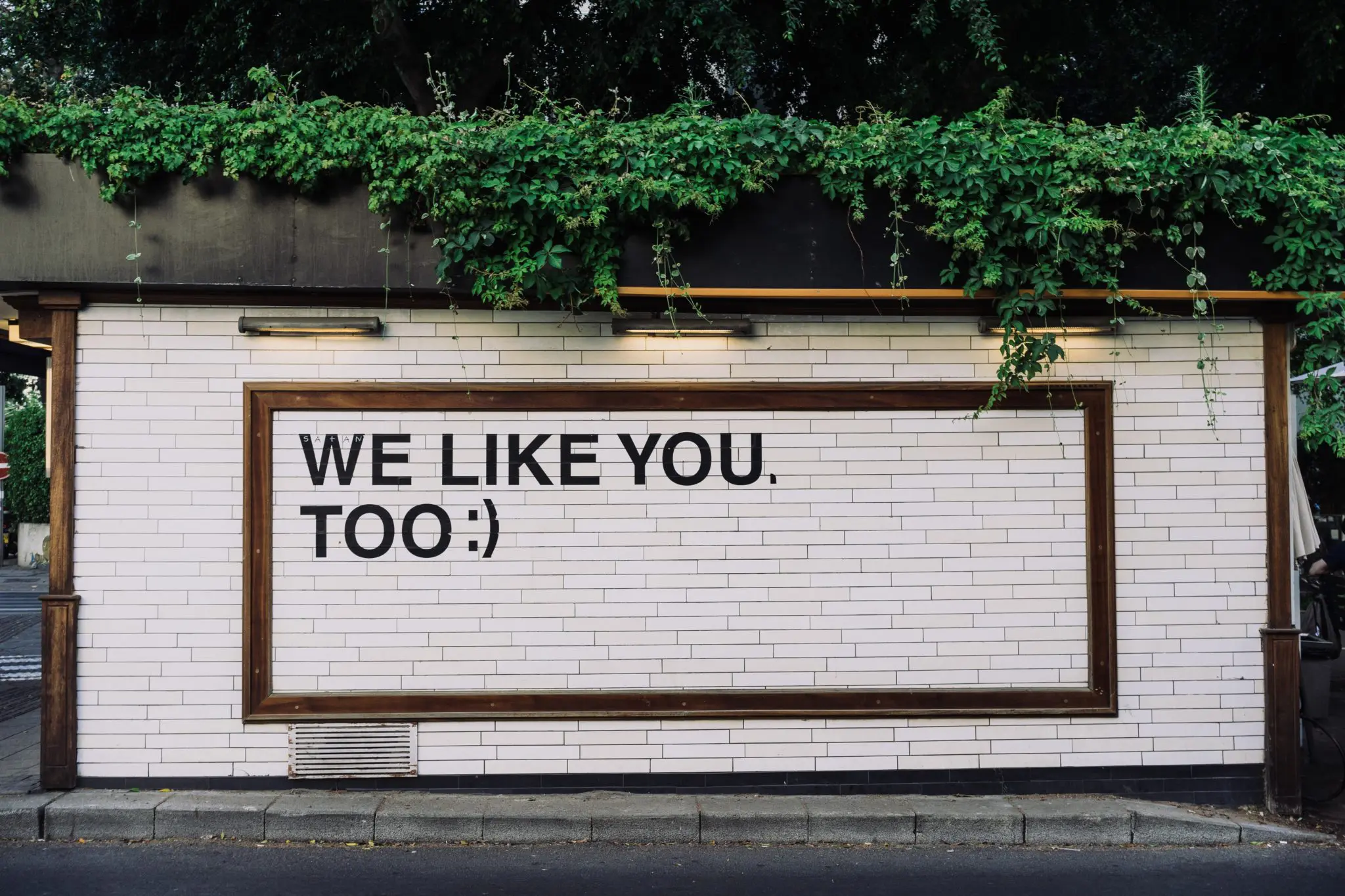 "We like you too :)" written on a white metro tile style wall outside
